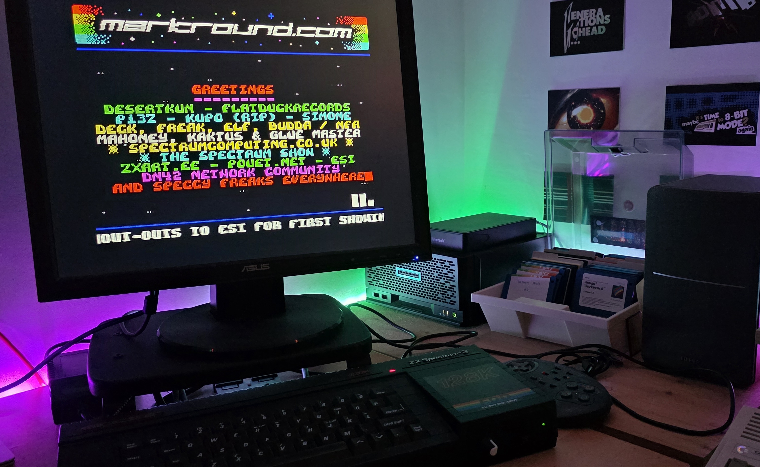 昏暗桌面上的 ZX Spectrum +3 实机：显示器里正运行刚做好的 intro，顶部是变色像素 logo，下面是 GREETINGS 名单和滚动文字，桌前摆着 Spectrum 128K 与键盘