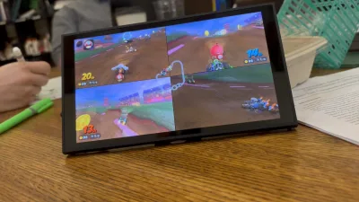 木桌上的 Switch 2 屏幕里跑着四人对战的 Mario Kart World，画面被分成四格，场上是泥地赛道