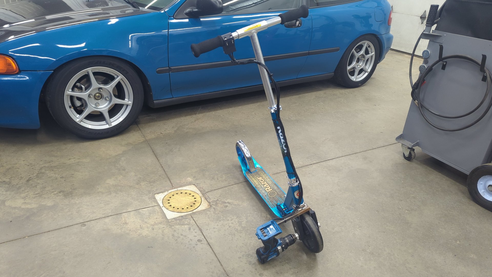 照片：一辆蓝色的 Razor 滑板车，前轮上装着一把蓝色无绳电钻，停在车库地面上，旁边是一辆蓝色的本田 Civic