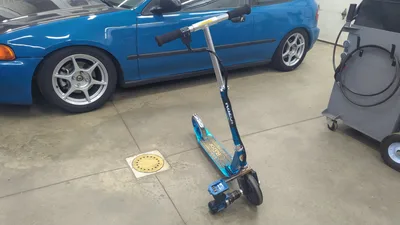 照片：一辆蓝色的 Razor 滑板车，前轮上装着一把蓝色无绳电钻，停在车库地面上，旁边是一辆蓝色的本田 Civic
